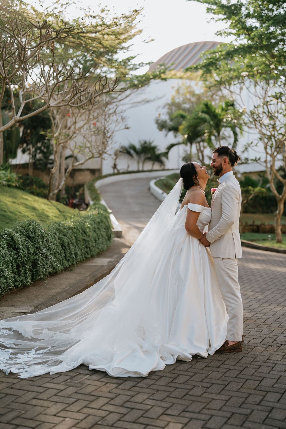 A wedding couple stands on a brick path with a flowing white dress and veil against a tropical backdrop.