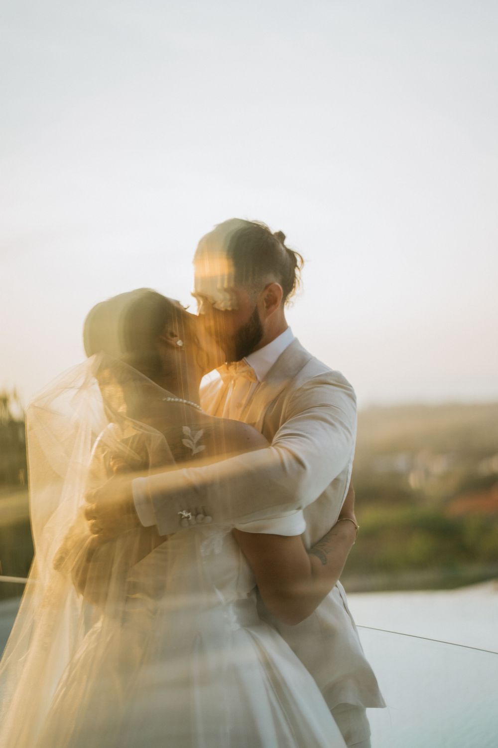 A romantic wedding couple embraces during golden hour sunset with soft light creating an ethereal glow through the veil.