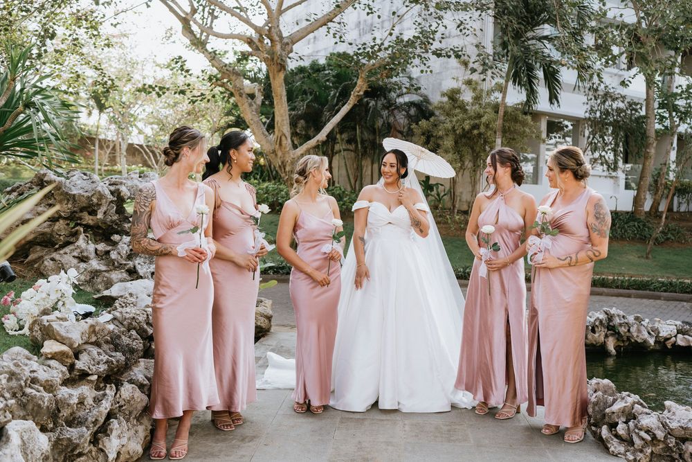 Bridesmaids in pink dresses gather around bride in garden setting.