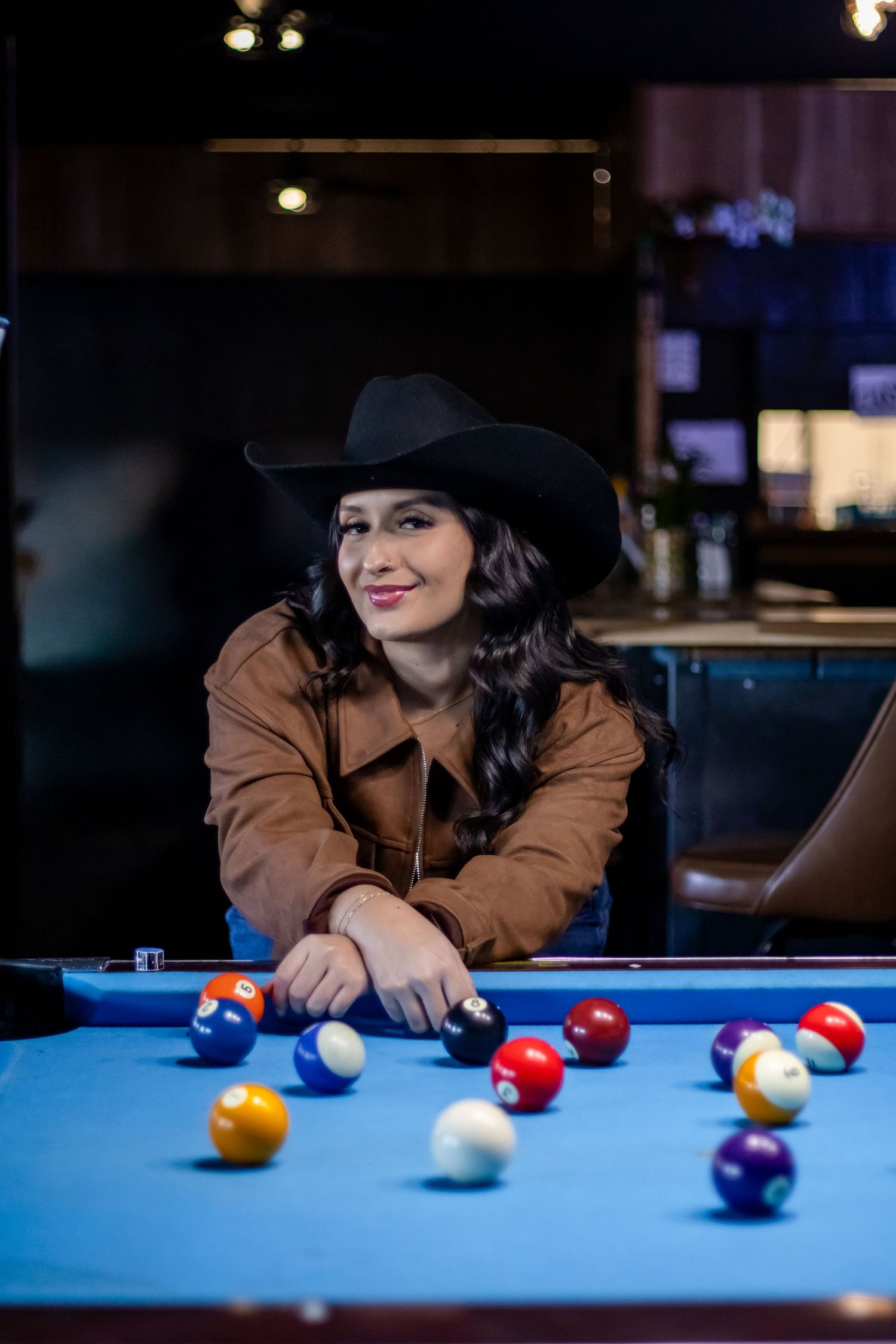 Woman in black cowboy hat smiling while leaning over blue pool table.