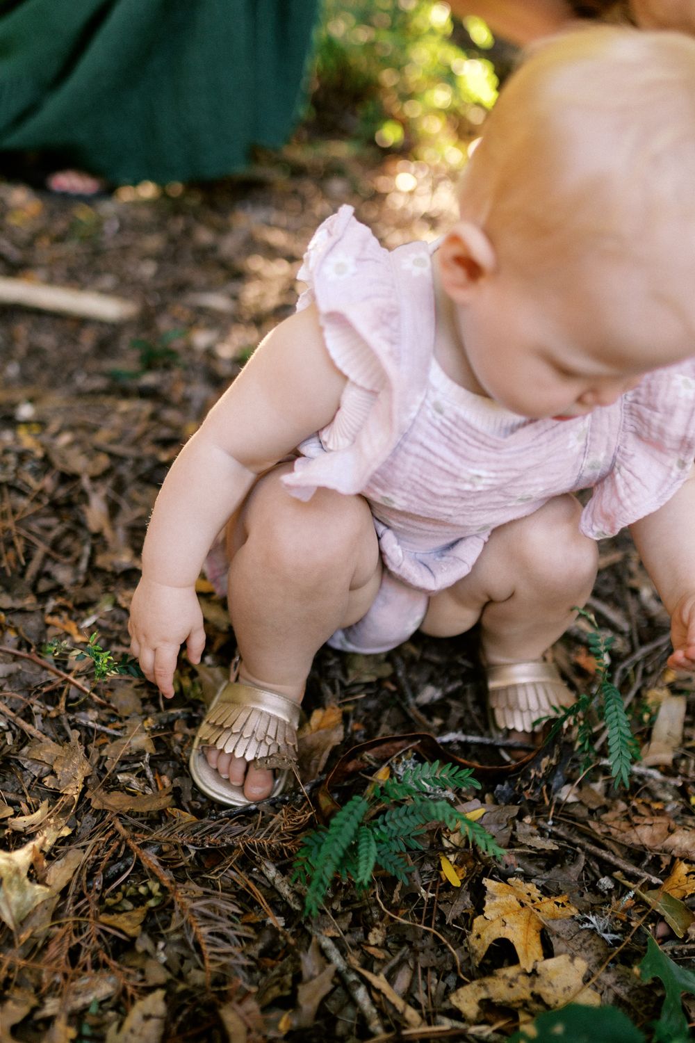 Baby wearing white dress and gold moccasins crawls on ground covered with fallen leaves.