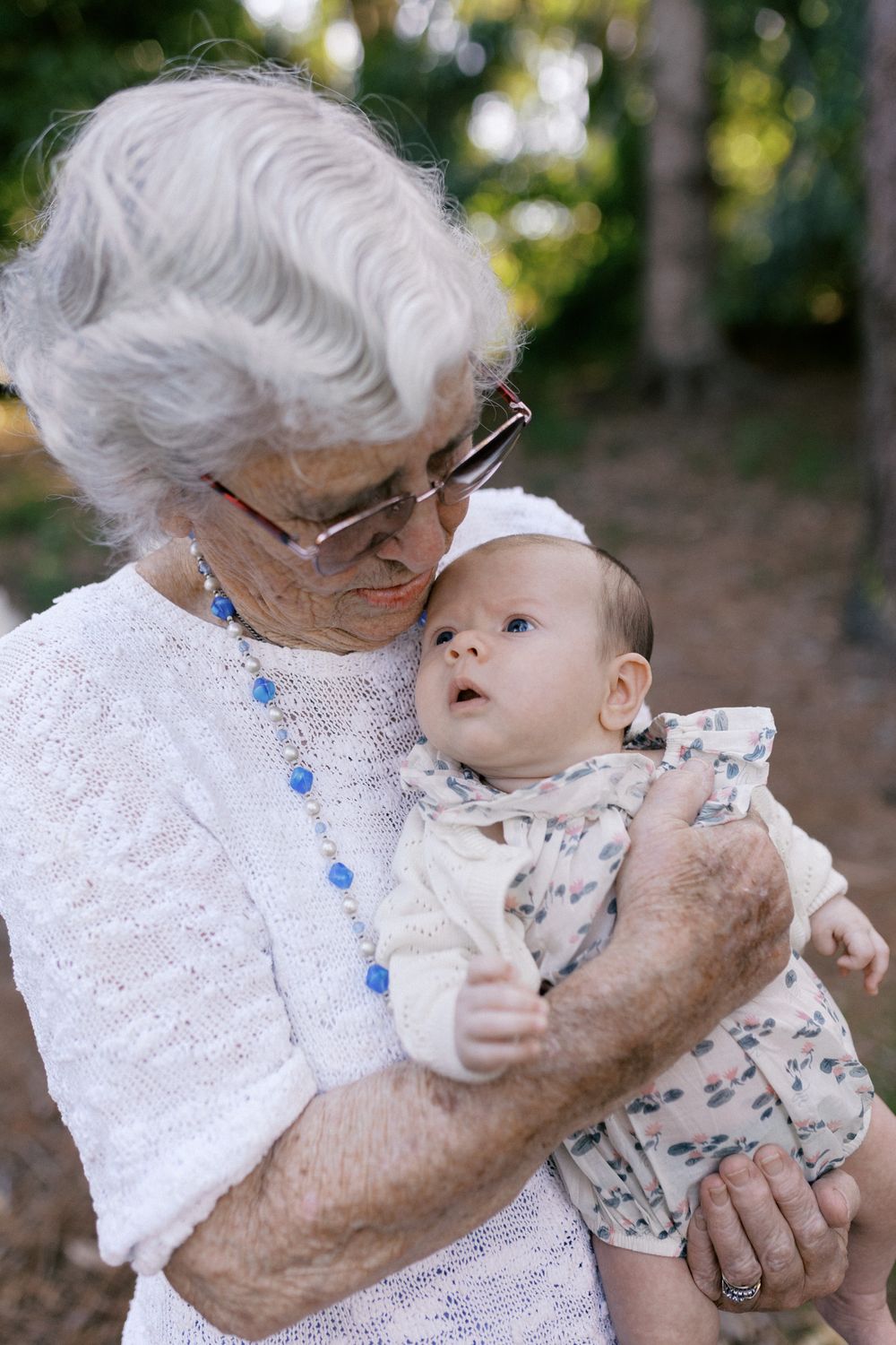 An elderly woman in white lace holds and kisses a baby in a tender outdoor moment.