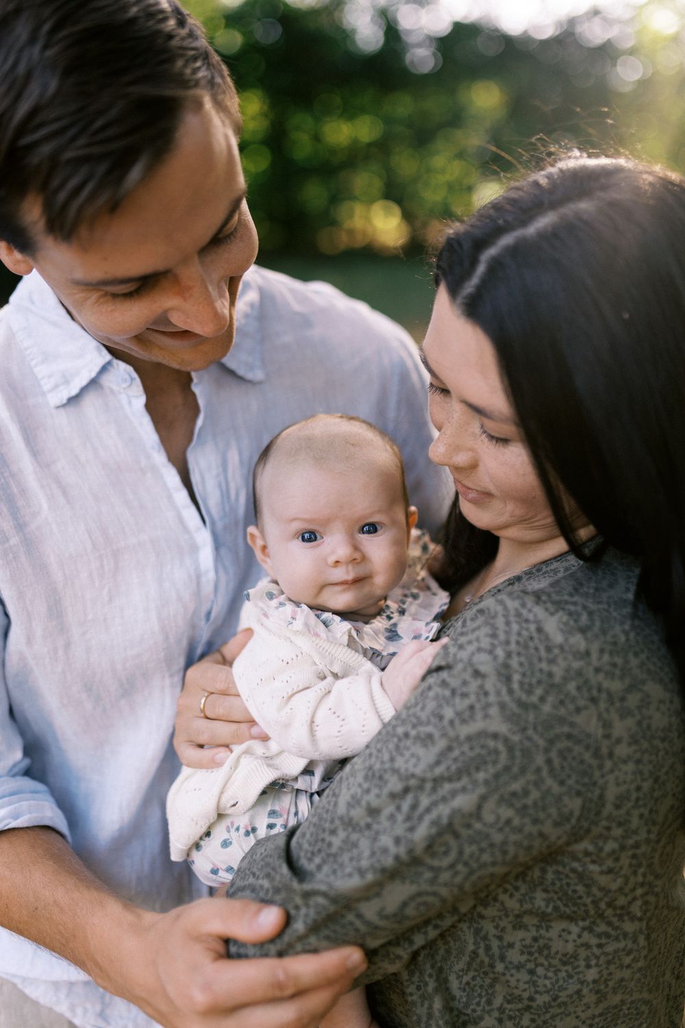 A tender family moment outdoors shows parents cuddling their baby in a warm embrace.