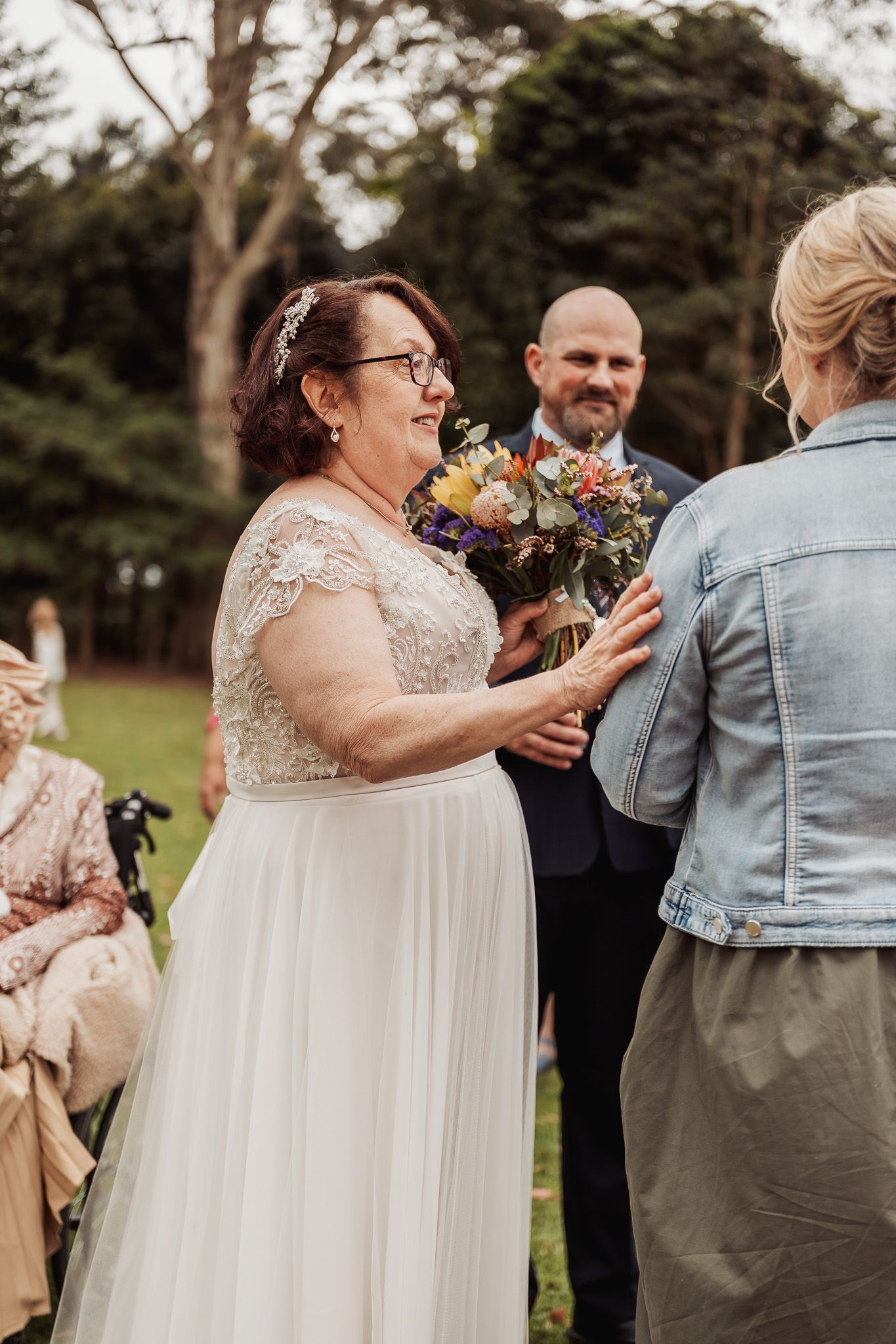 Wedding ceremony taking place outdoors on a red carpet aisle with guests seated on white chairs in a garden setting.