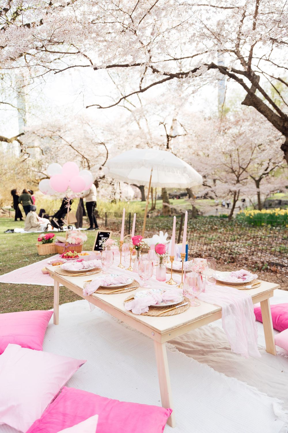 Pink themed outdoor picnic setup under blooming cherry blossoms with elegant table settings and cushioned seating.