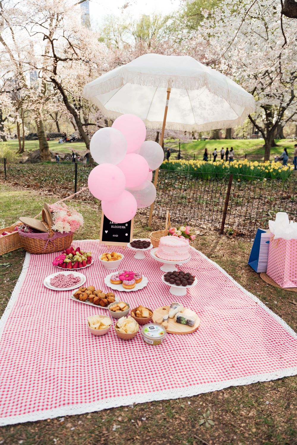 Picnic setup with pink gingham blanket, white umbrella, and balloon decorations under blooming cherry blossom trees.