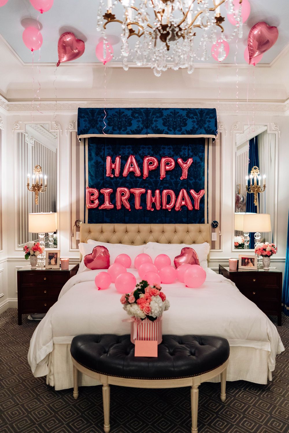 Luxury hotel room decorated for a birthday celebration with pink balloons, heart shapes, and 'Happy Birthday' text on the wall.