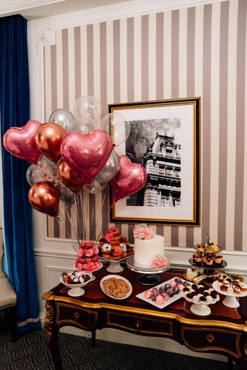 Elegant dessert table display with pink and red heart balloons, cakes, and treats against a striped wall with blue curtains.