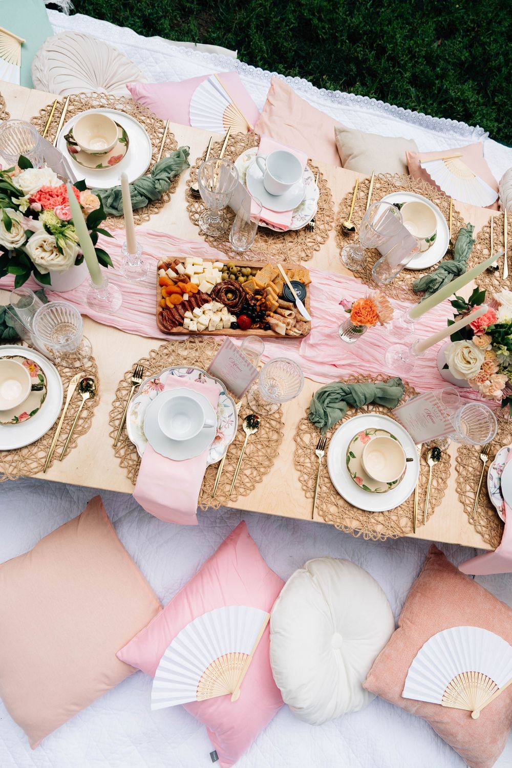 Elegant outdoor picnic setting with pink and white decor, teacups, decorative fans, and plush cushions arranged on blanket.