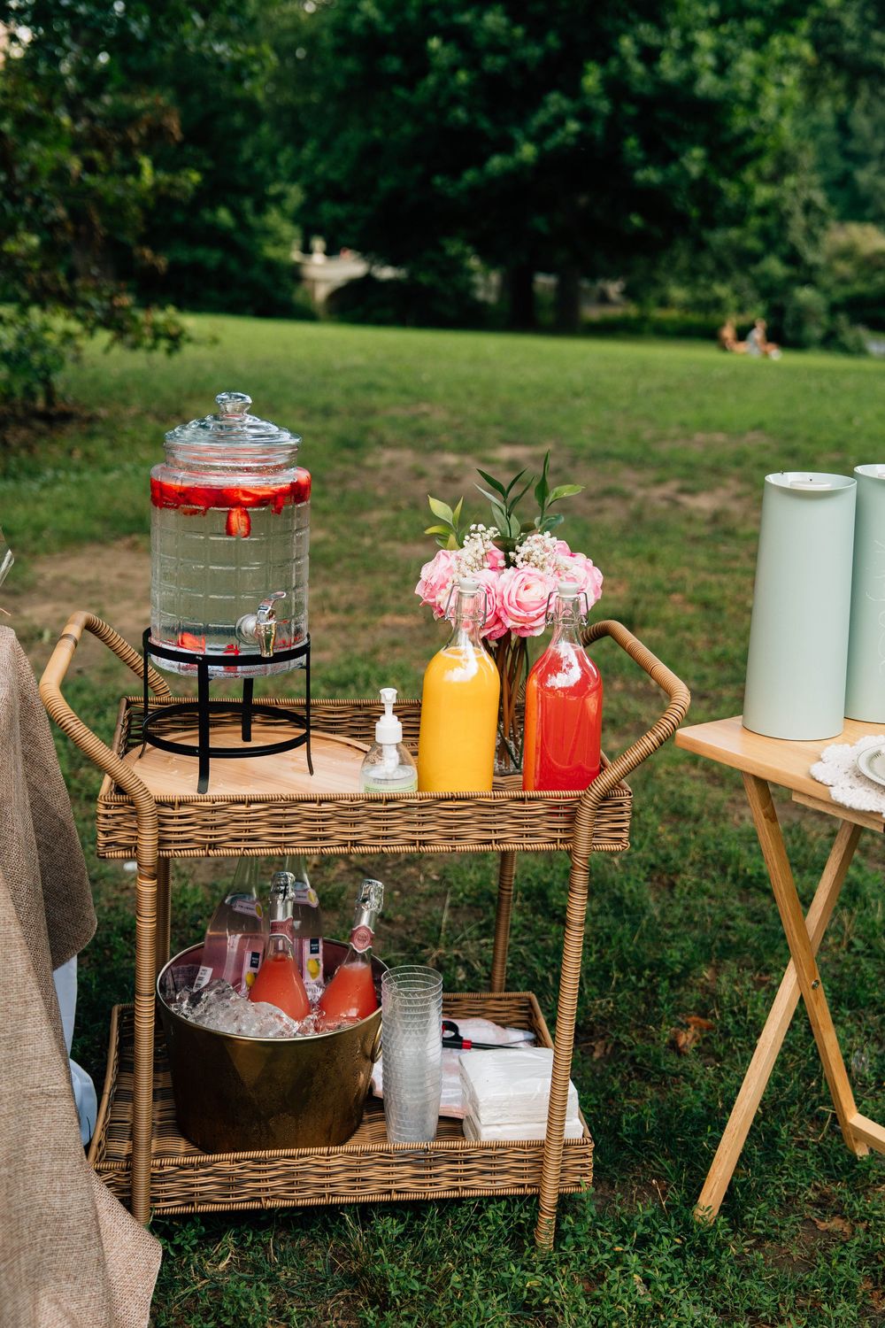 Rustic outdoor beverage station with wicker bar carts displaying drinks and glassware in a garden setting.