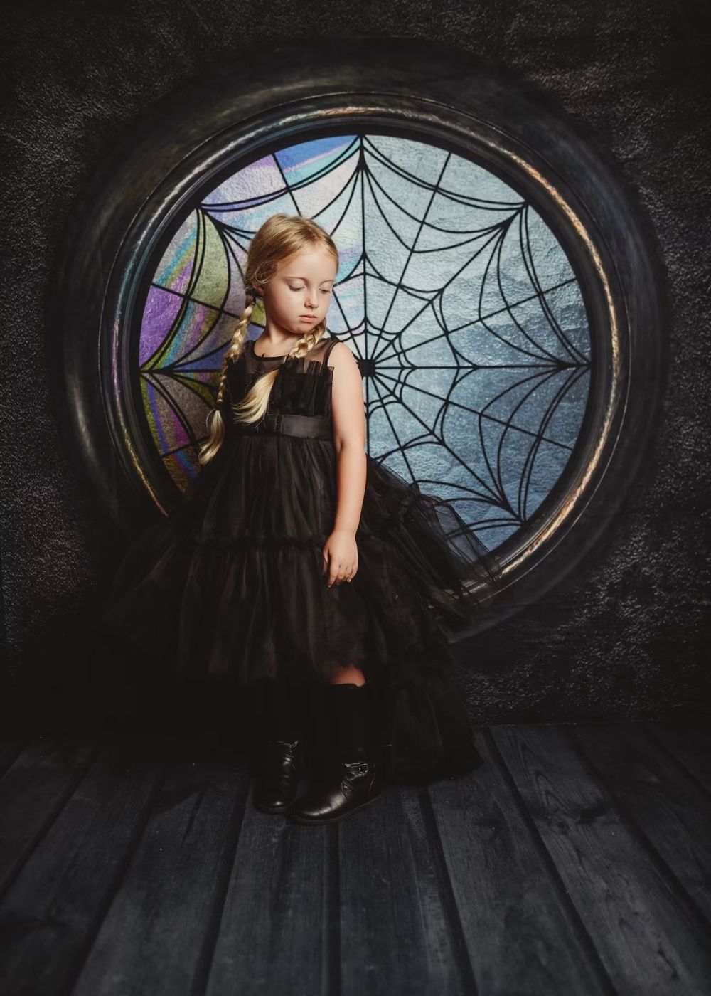 Young girl in black dress and boots poses before a gothic spider web stained glass window.