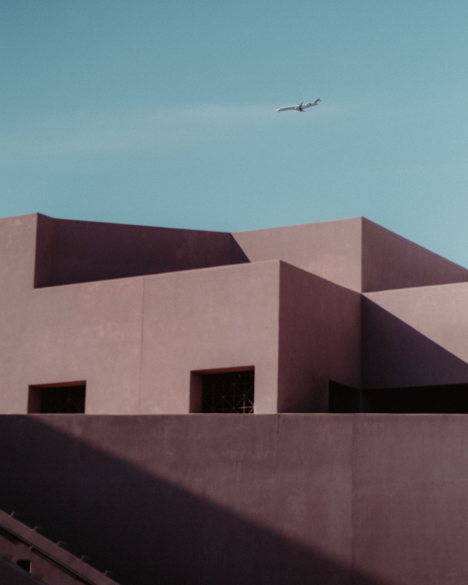 Minimalist architecture with geometric shapes and an airplane flying overhead against blue sky.