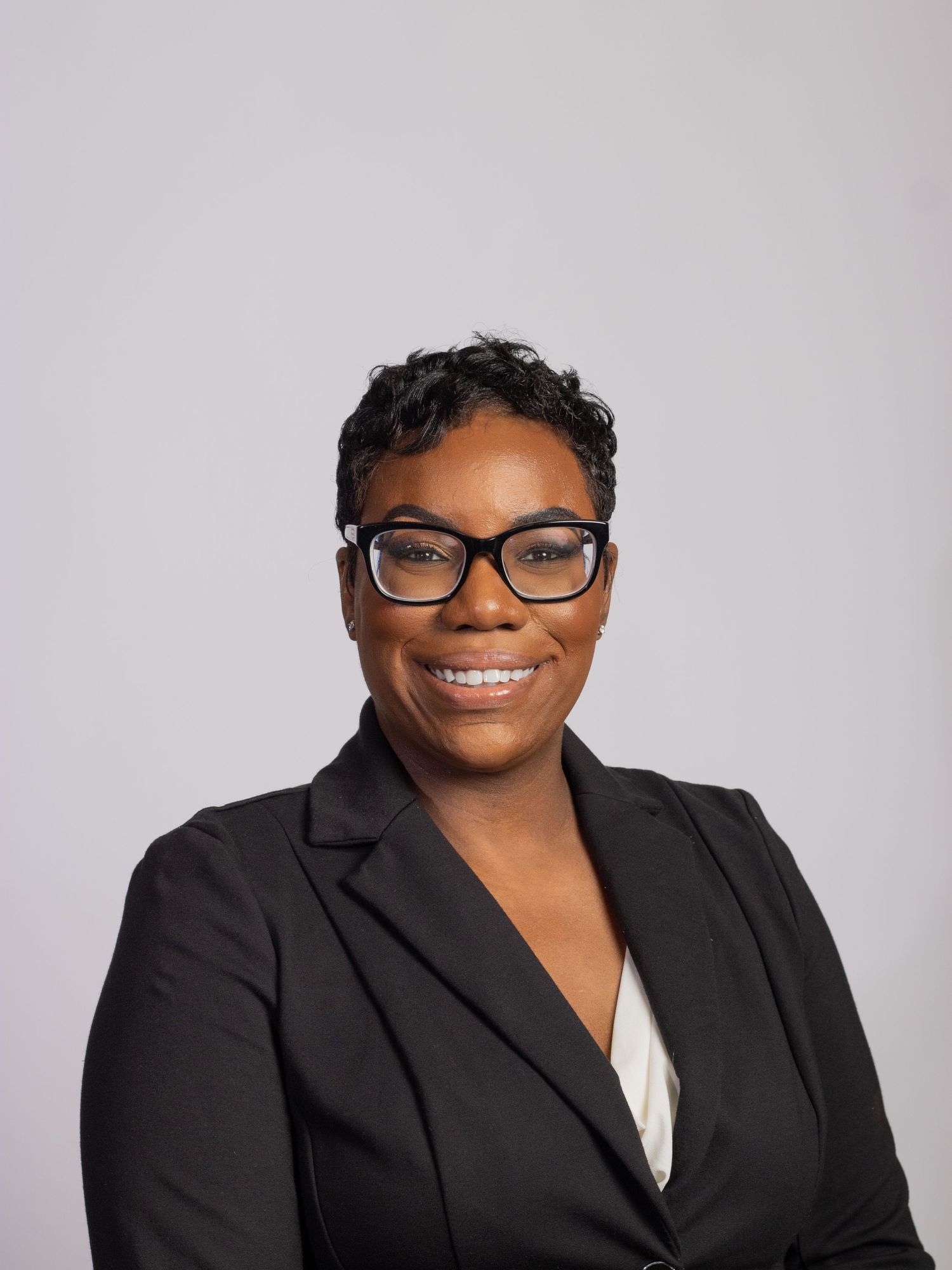 Professional woman in black blazer and glasses smiles in corporate headshot photo.