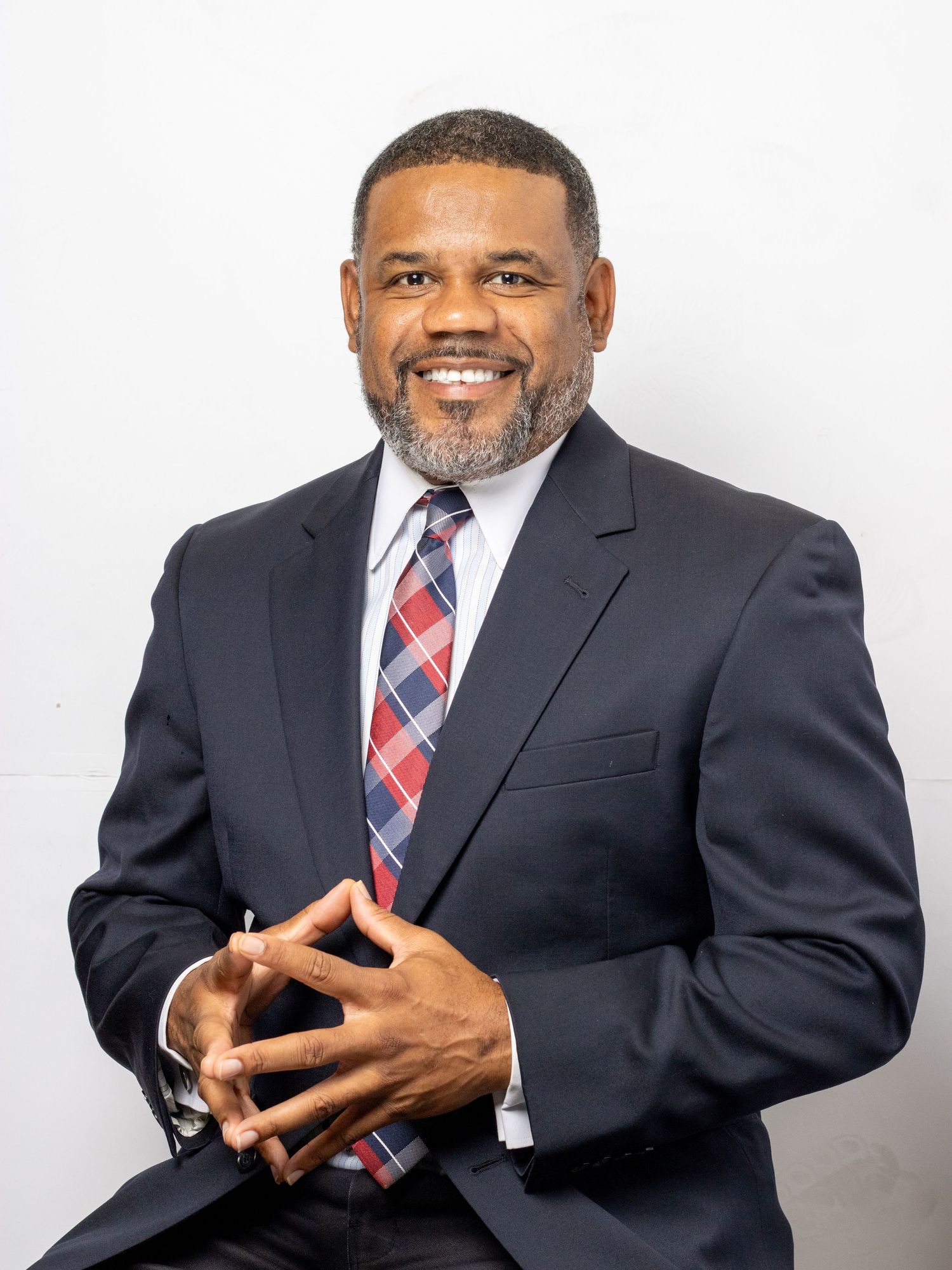 Professional man in navy suit and plaid tie smiling with hands clasped against a white background.