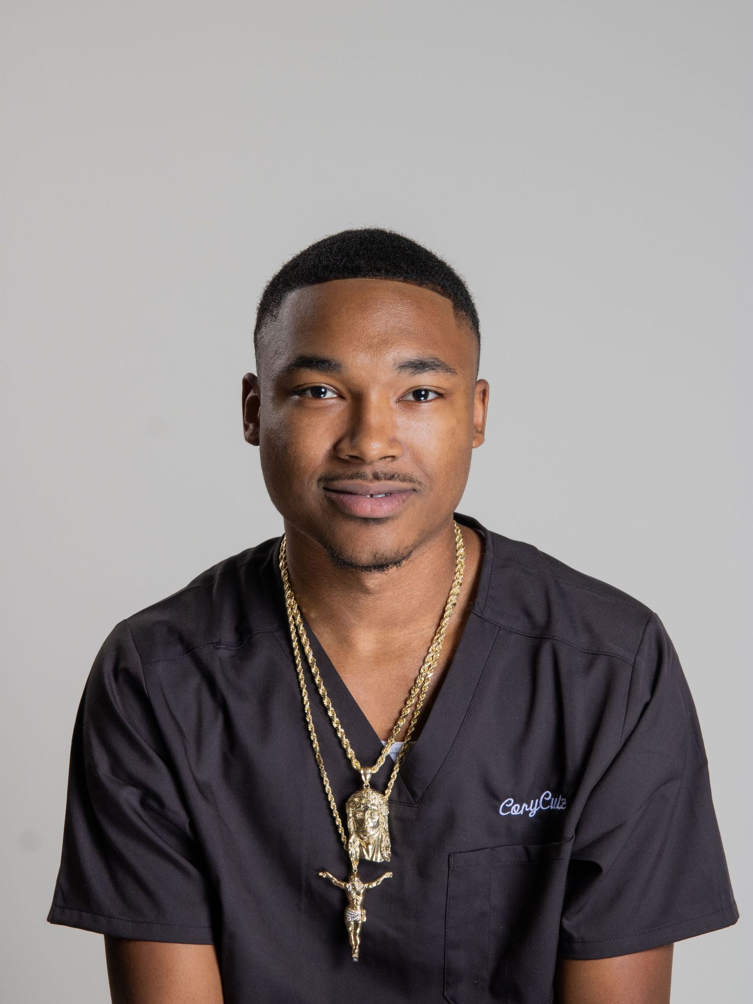 Young man in dark scrubs wearing gold chain necklaces posing for a professional headshot.