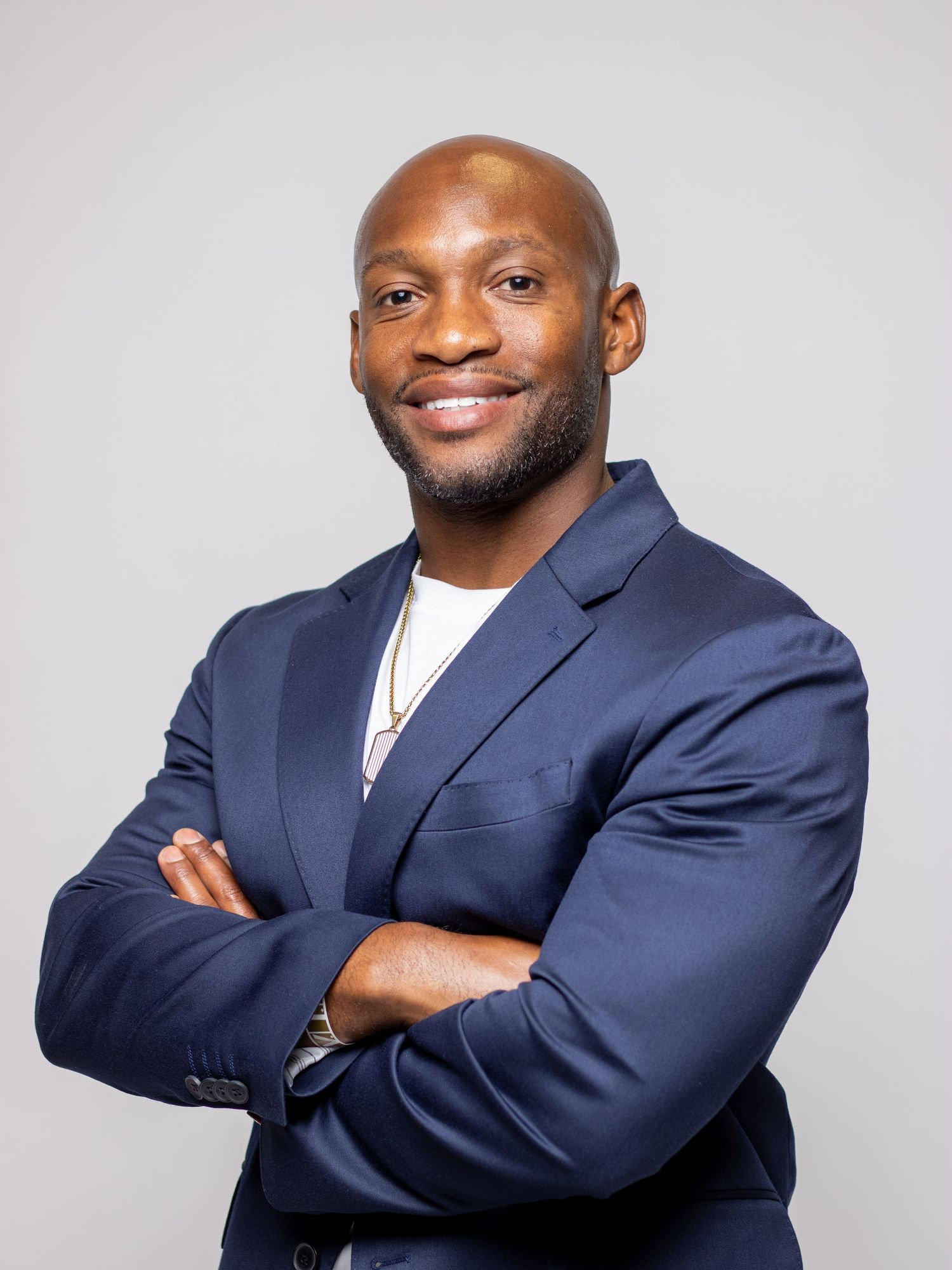 Confident Black man in navy blazer with arms crossed smiling against gray background.