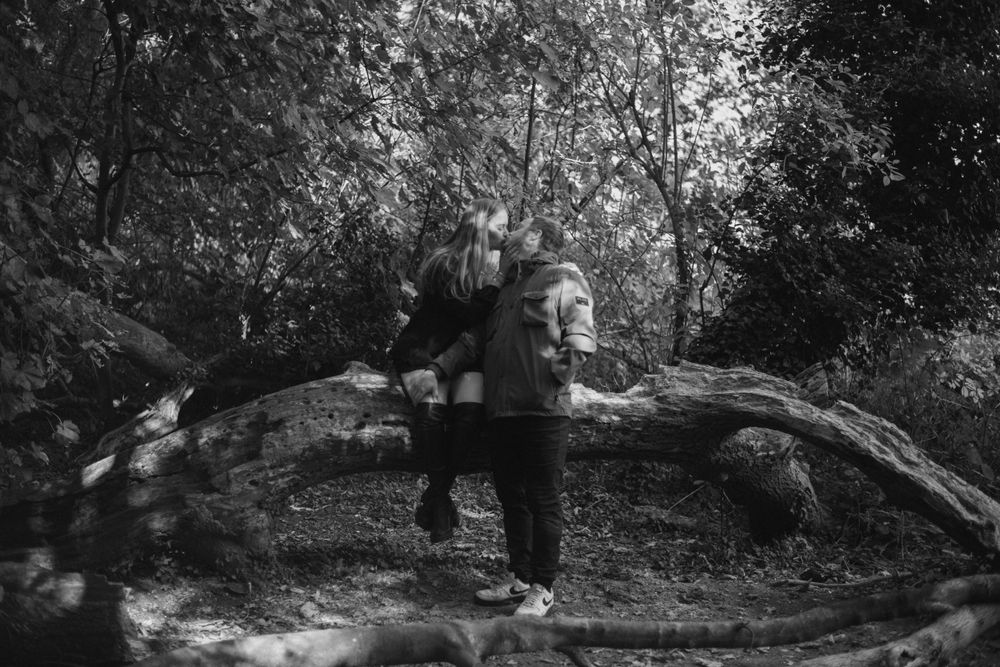 A couple stands together in front of a fallen tree branch in a moody black and white forest setting.