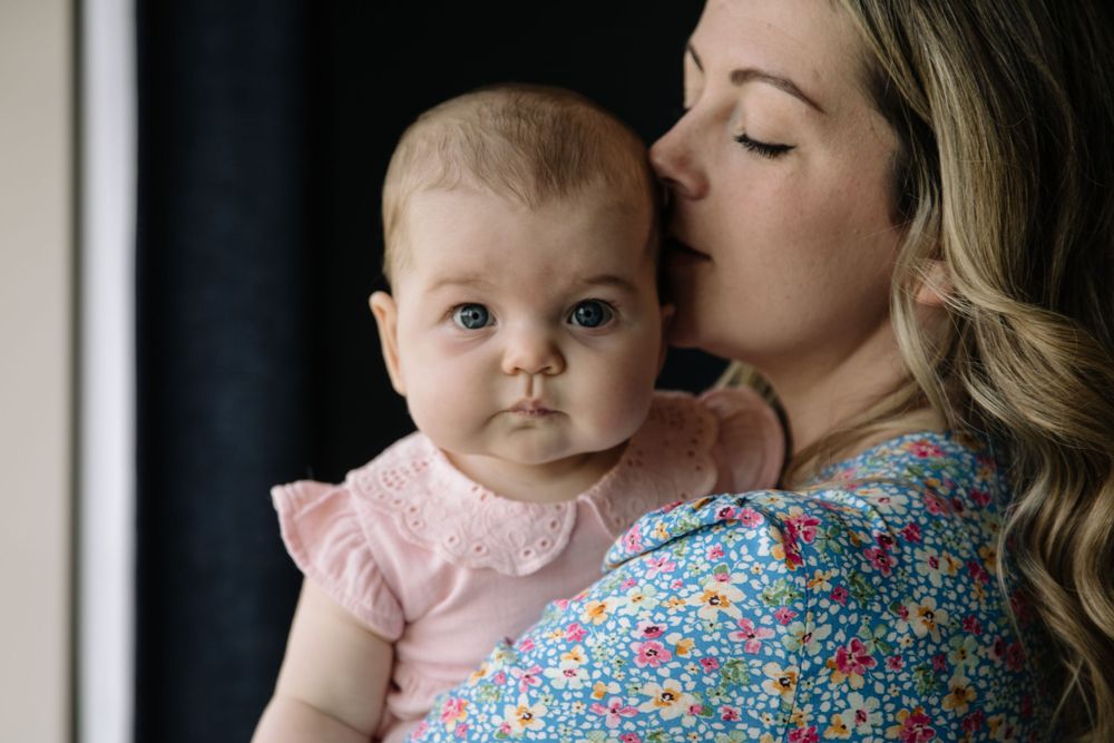 Mother in floral dress kissing baby in pink outfit against dark background.