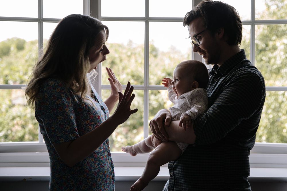 A candid family moment by a large window as parents interact with their infant.
