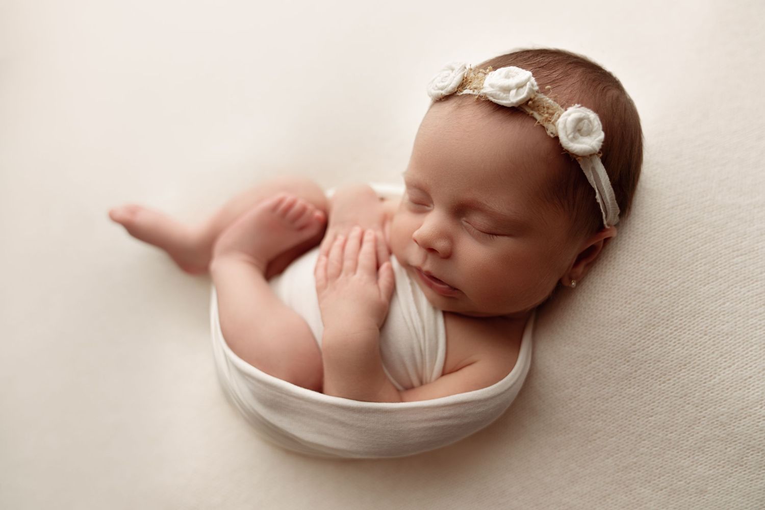 Sleeping newborn in white wrap and floral headband posed in cream bowl.