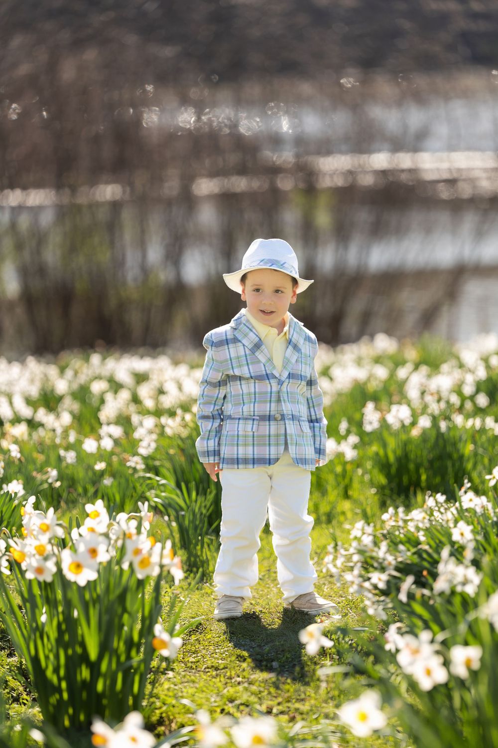 A young boy in a white hat and plaid blazer stands confidently among white daffodils in a sunny spring field.