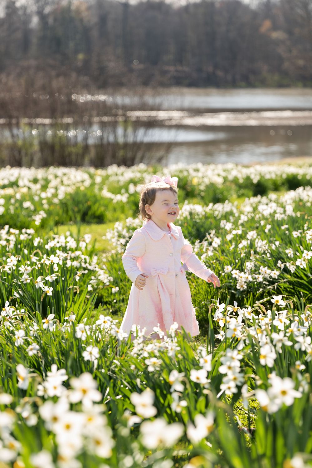 A joyful toddler girl in a pink dress laughs while running through a sunlit field of white and yellow daffodils.