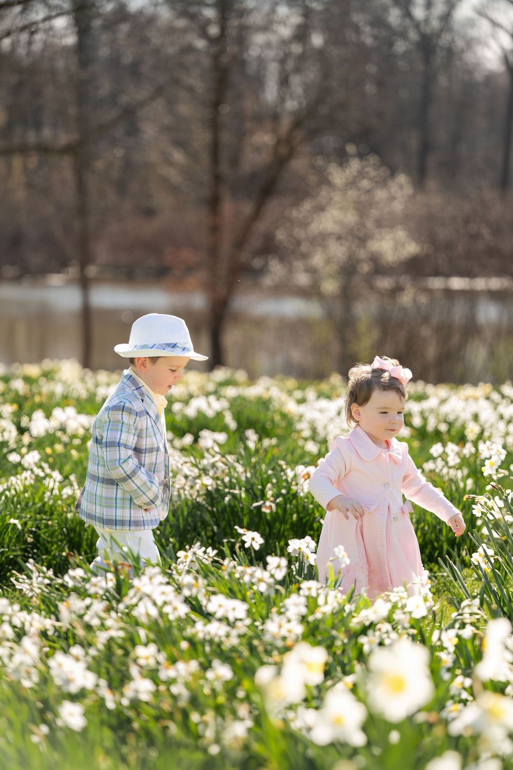 Two young children, boy in plaid and hat, girl in pink dress, playing in daffodil field.