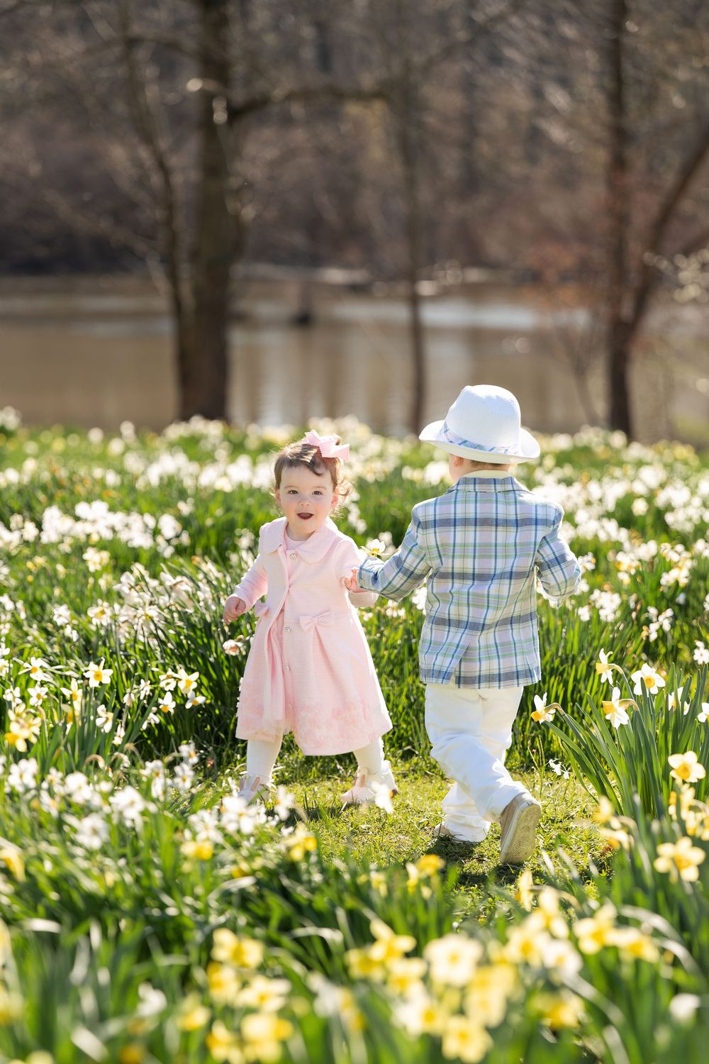 Two young children holding hands and laughing together in a sunny field of spring daffodils.