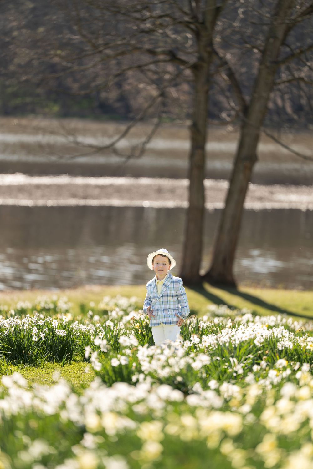 Young boy in white hat and plaid shirt standing alone in a sunny daffodil field near a lake.