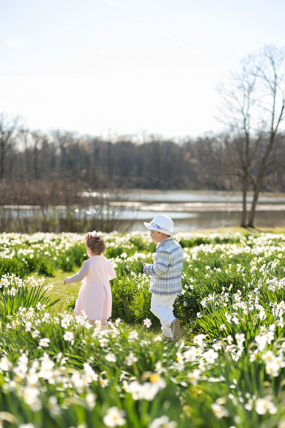 A young girl in a pink dress and a boy in a plaid jacket walk together through a field of white daffodils.
