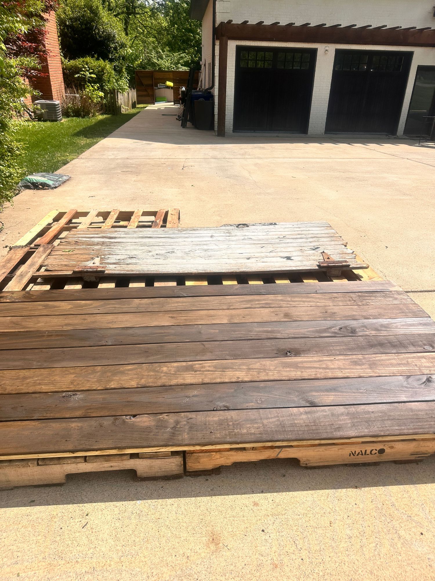 Wooden shipping pallets stacked on a concrete driveway near a two-car garage surrounded by green trees.