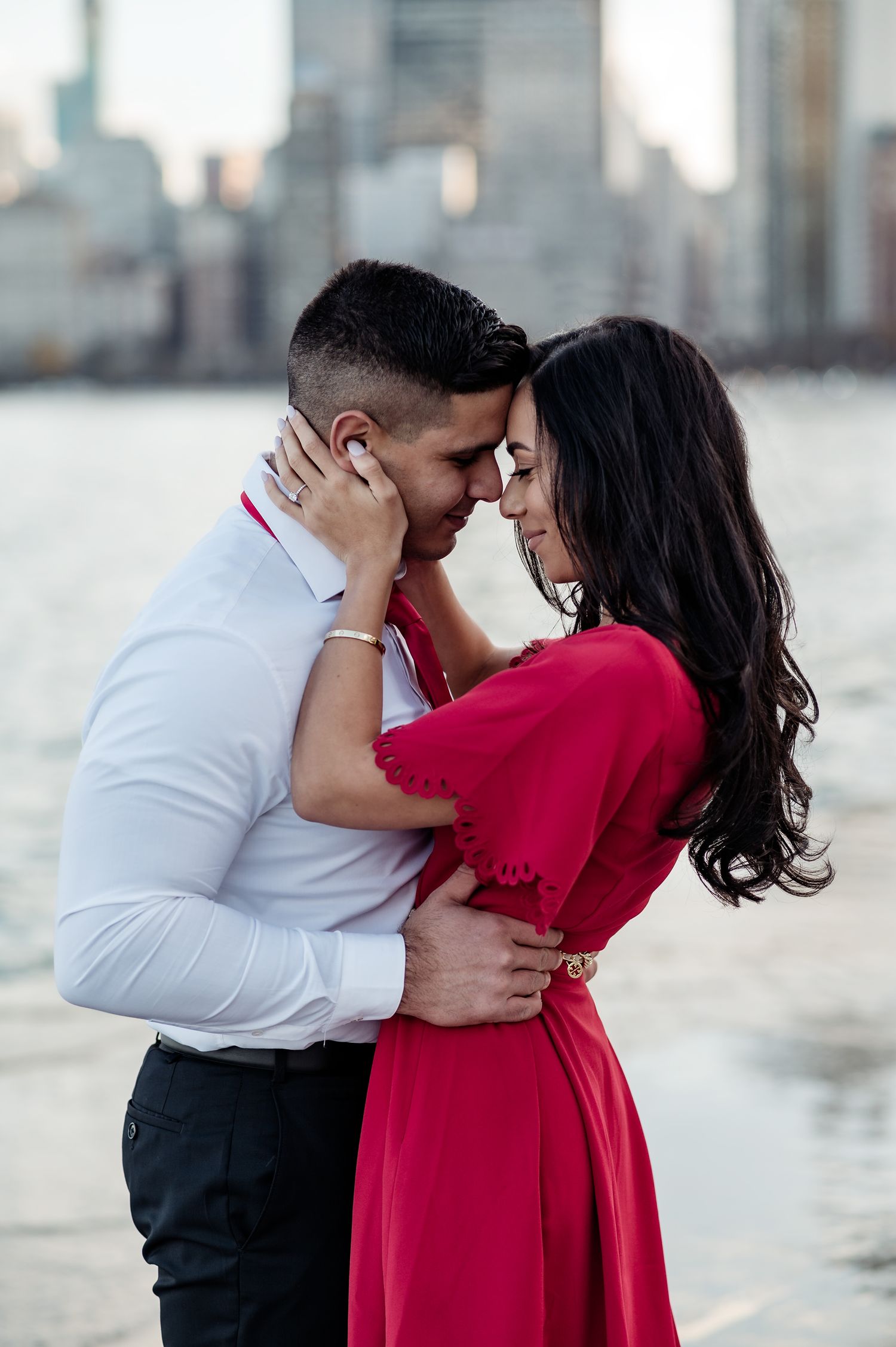 Romantic couple embracing near waterfront with city skyline, woman in red dress and man in white shirt touching foreheads.