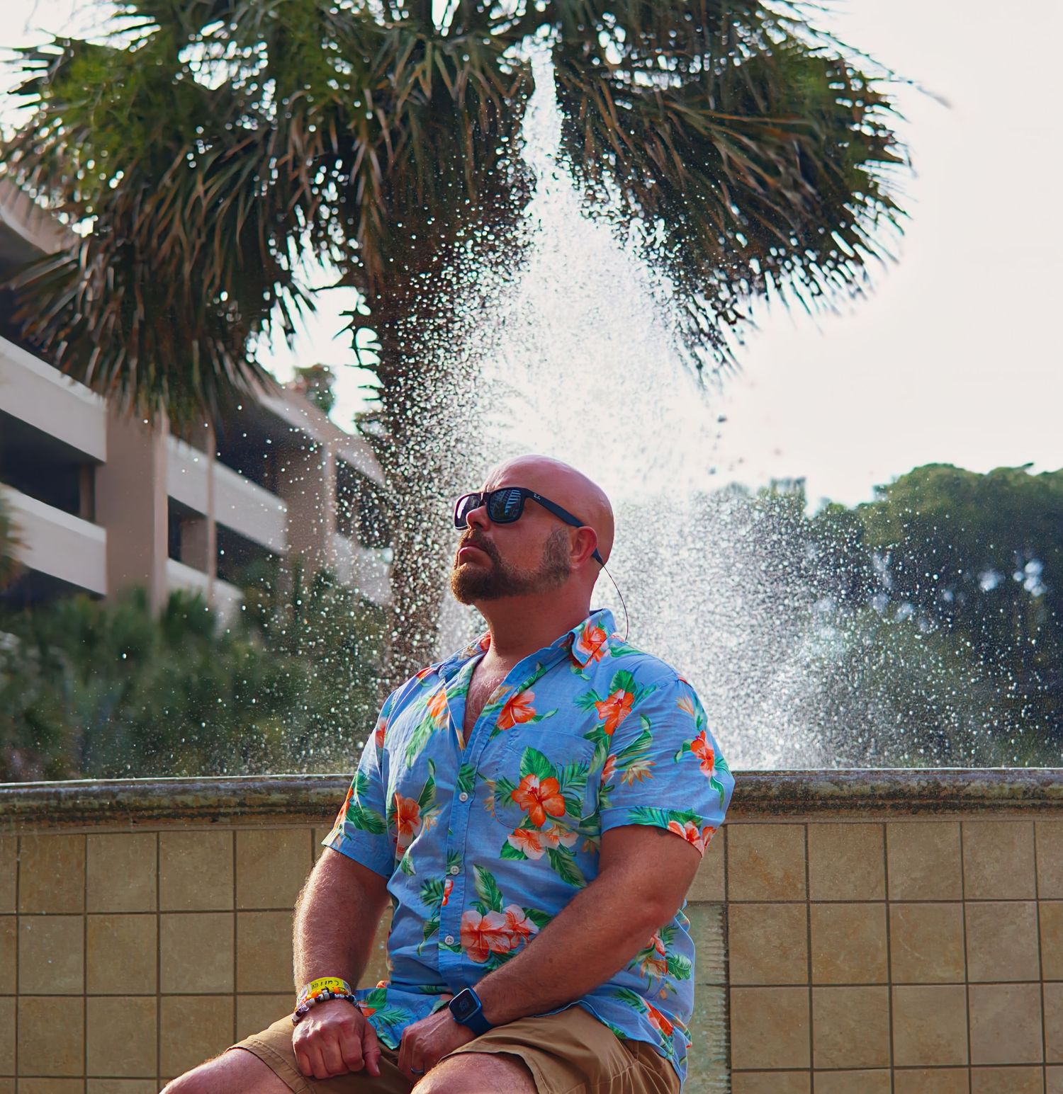Bald man in colorful floral Hawaiian shirt sitting by a fountain with palm trees in background.