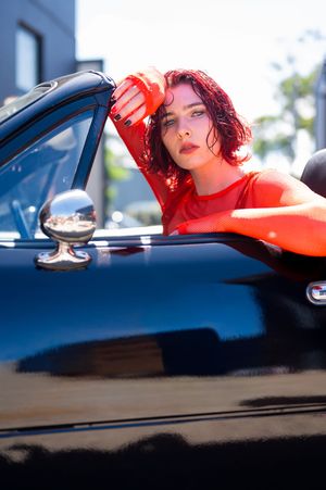 A woman with red hair in a red outfit leans out of a classic black convertible car on a sunny day.