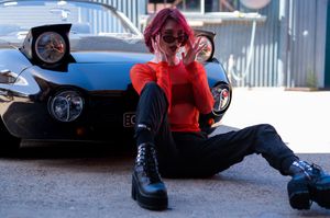Woman with pink hair in red top and black pants sits in front of a black sports car wearing platform boots.