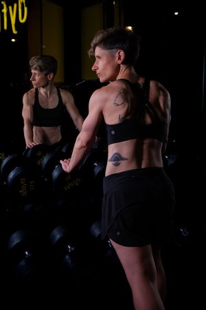 Muscular woman with tattoos in black sports bra and shorts lifting dumbbells in a dark gym, reflected in mirror.
