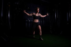 Woman performing cable fly exercise in dark gym wearing black sports bra and shorts, arms extended wide.