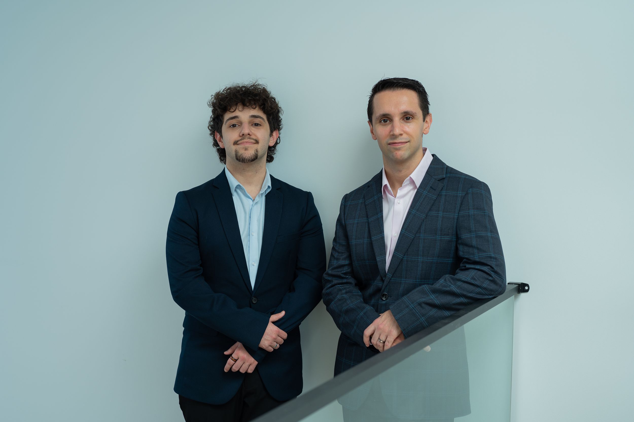 Two professional men in blazers posing together against a light blue wall in a modern office setting.