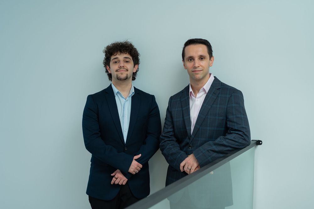 Two professional men in blazers posing together against a light blue wall in a modern office setting.