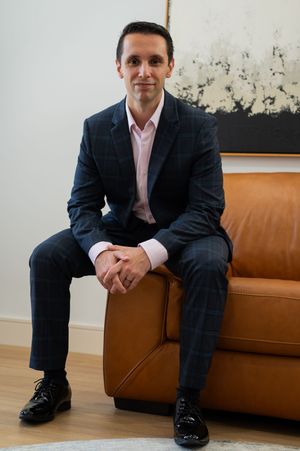 Professional man in navy plaid suit sitting on brown leather sofa in modern office setting.