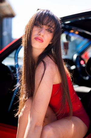 A young woman with long brown hair in a red dress poses beside a red car in bright sunlight.