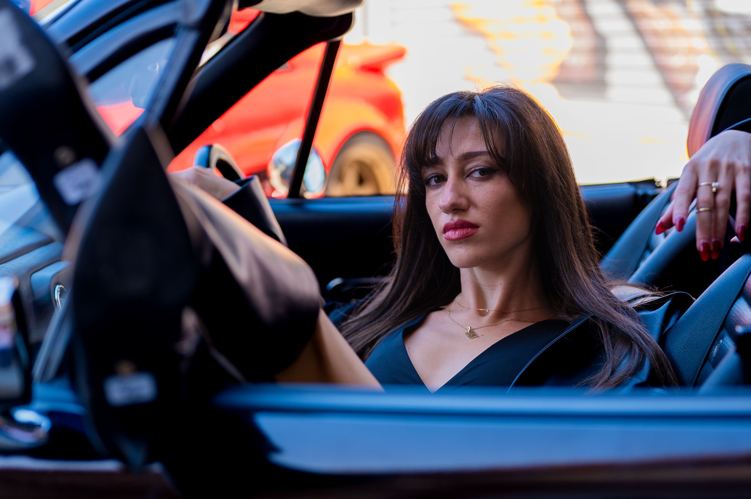 A stylish woman with long brown hair sits in a luxury convertible car, wearing a black dress.