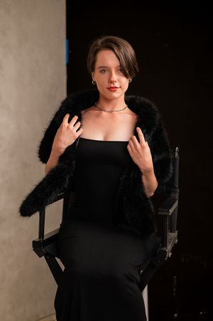 Woman in black dress and fur coat poses elegantly in a director's chair during a professional photo shoot.