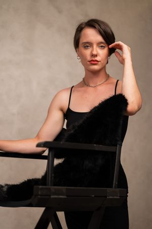 Woman in black spaghetti strap dress with fur stole sits in director's chair wearing silver necklace and earrings.