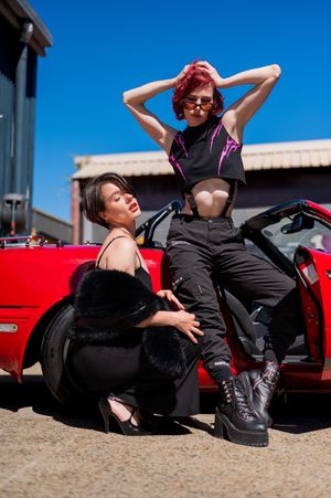 Two stylish women posing beside a red convertible sports car on a sunny day, wearing edgy black outfits.