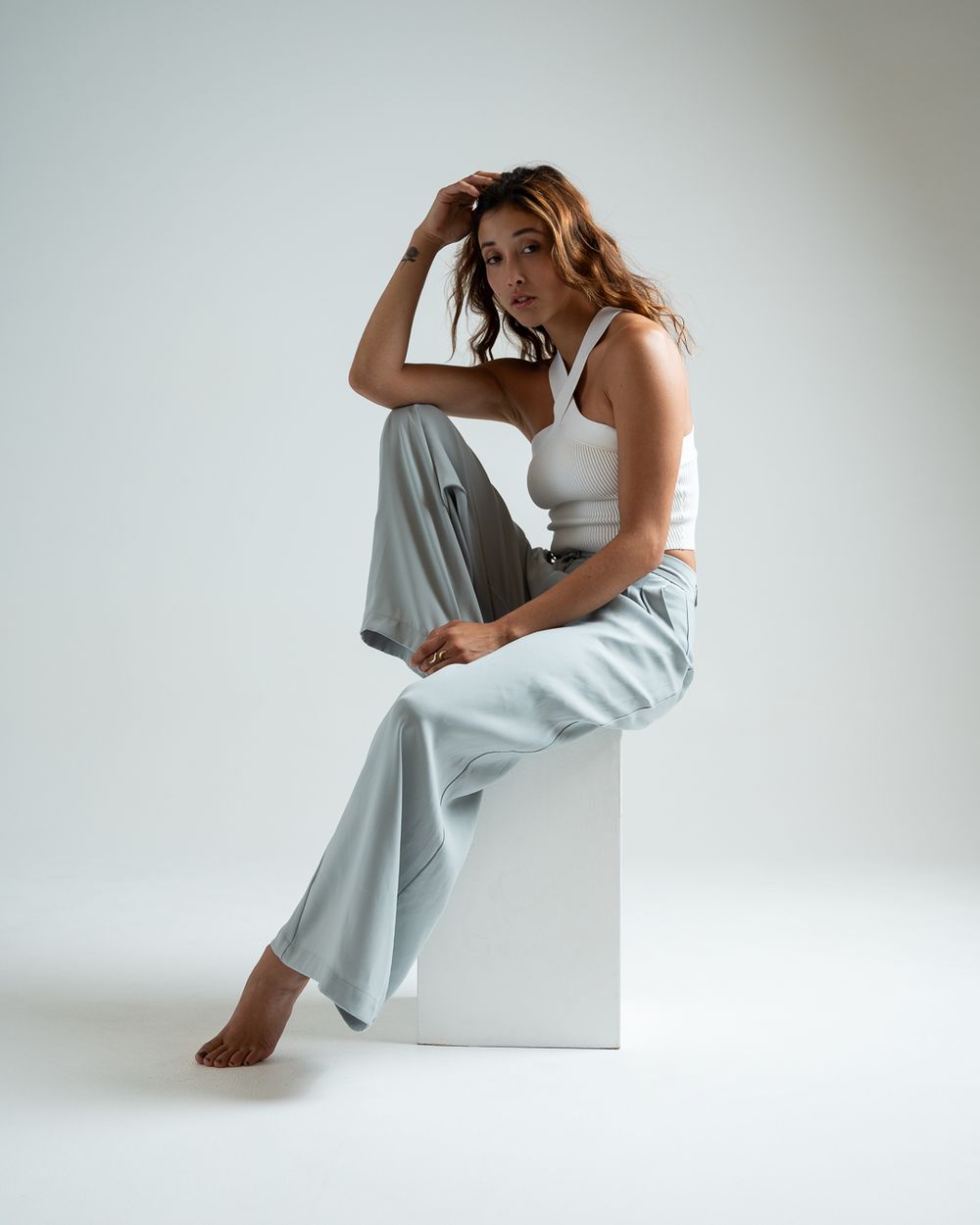 Woman in white crop top and light gray wide-leg pants sits barefoot on white cube against neutral background.
