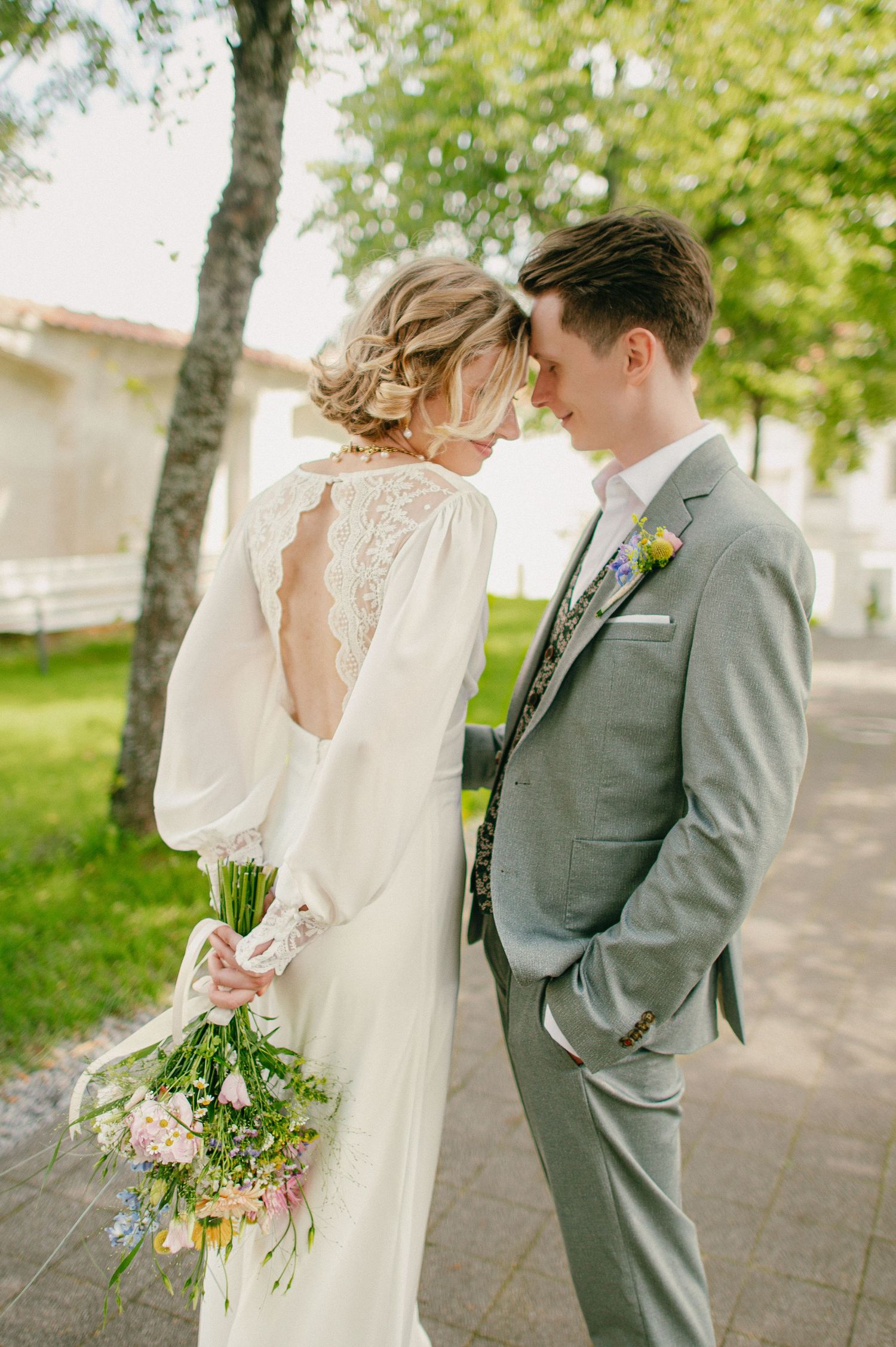 Bride in lace-back white gown holds wildflower bouquet as couple touches foreheads tenderly outdoors.