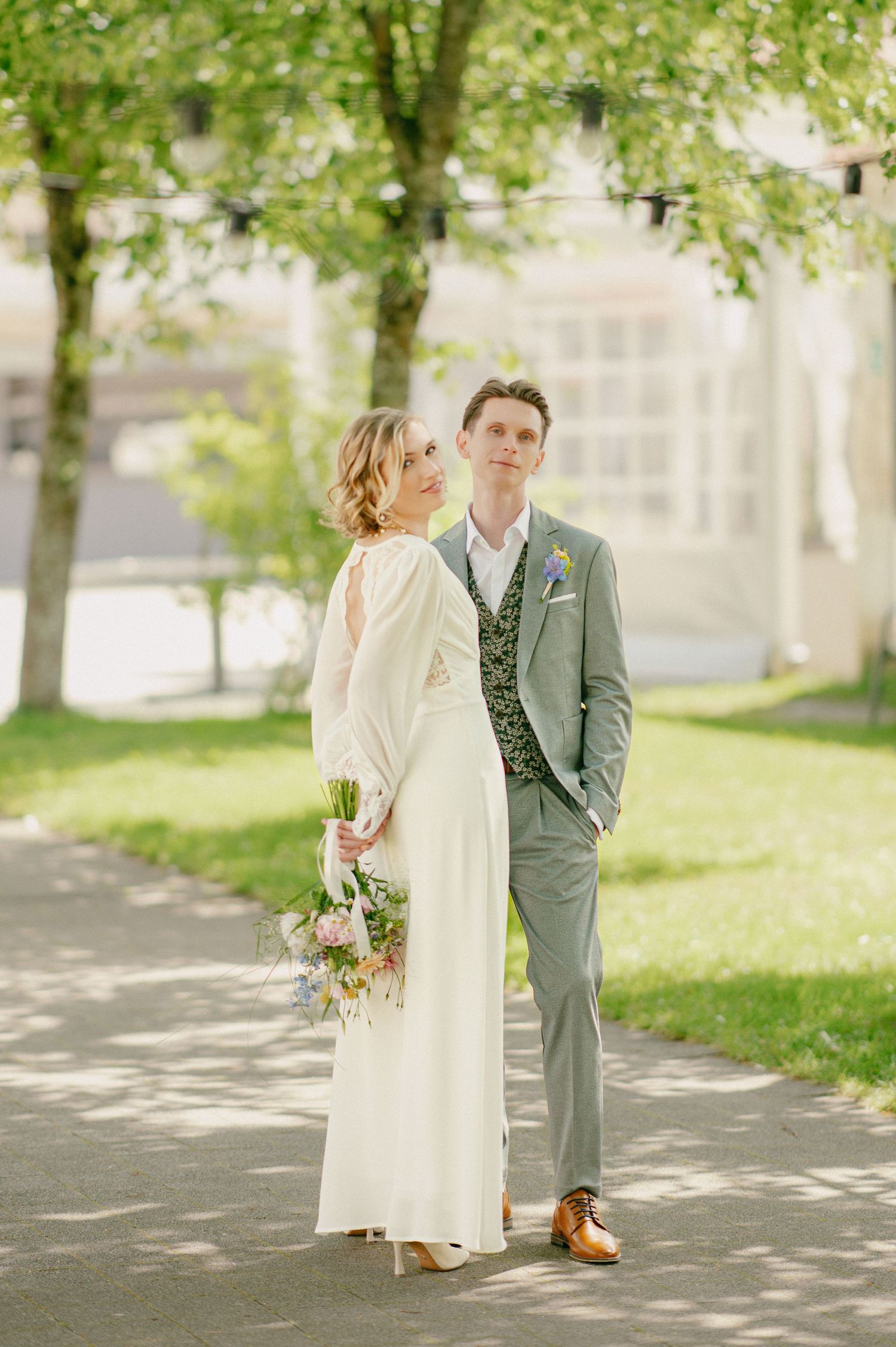 Elegant bride in a long white dress holds a bouquet while groom in gray suit and floral vest poses outdoors.