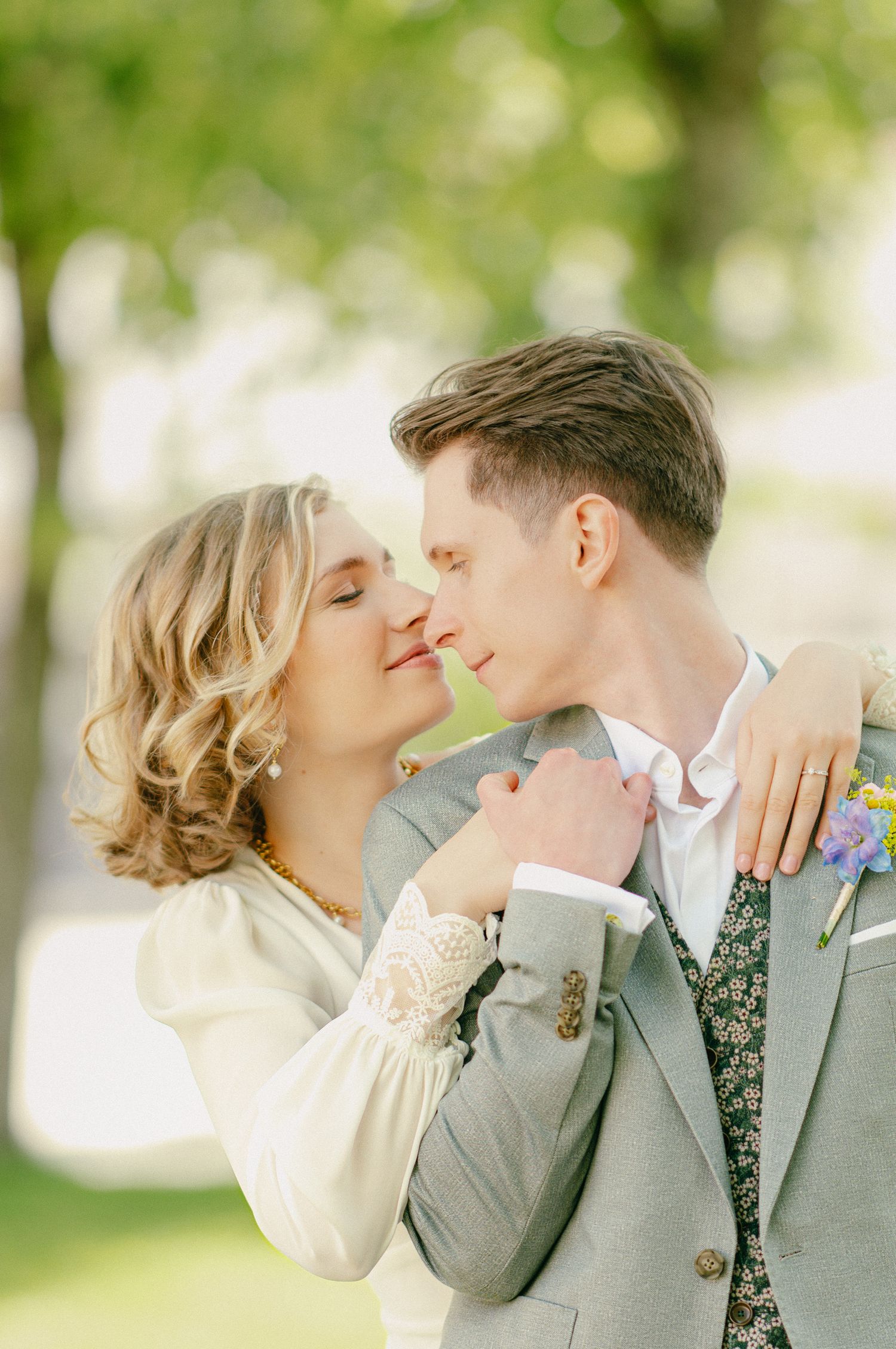 Bride and groom share a tender almost-kiss, she in a white lace dress, he in a gray suit with floral vest.