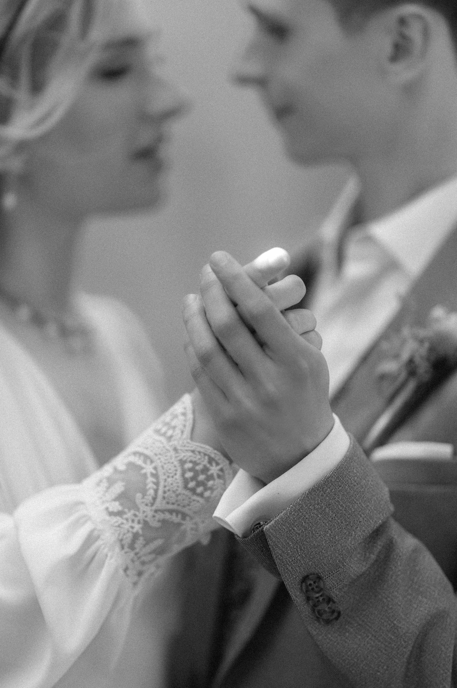 Black and white photo of bride and groom holding hands, wedding rings visible, sharing an intimate moment.